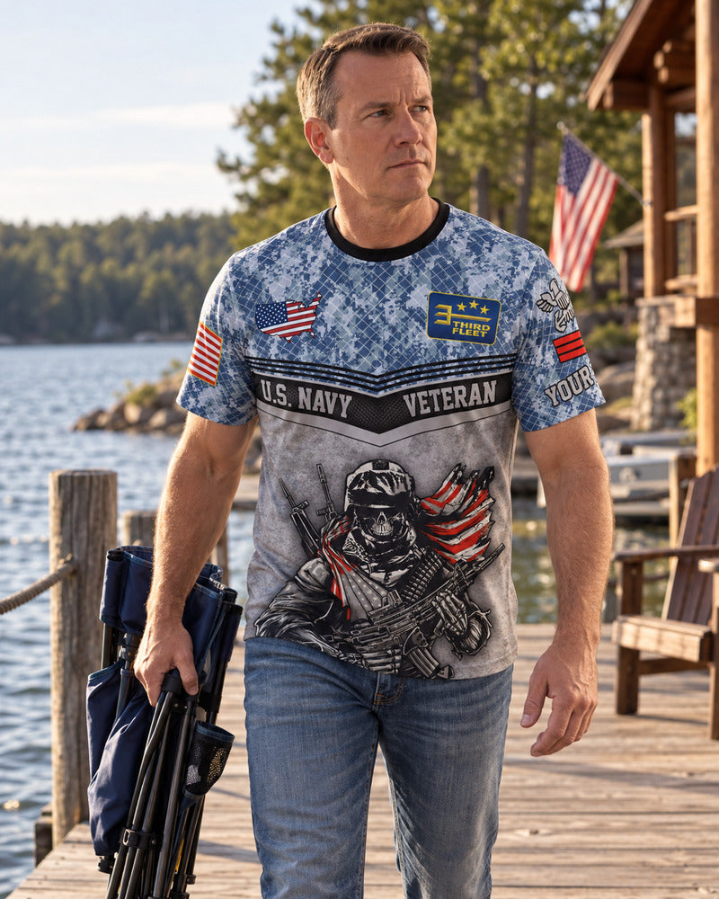 A man carrying a folded camp chair beside a lake in a Navy T-shirt.